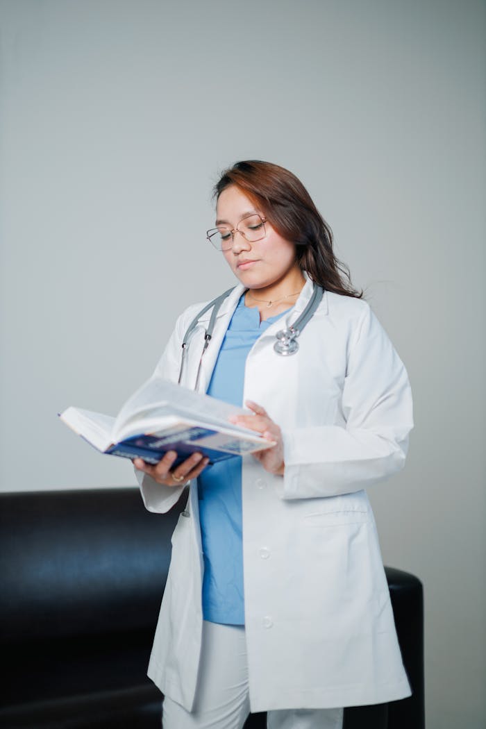 Female doctor in white coat and stethoscope reading a medical book indoors.